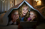 Young happy caucasian mother using a digital tablet with her little daughters before bedtime at home. Cute comfy siblings watching a movie on a digital tablet with their mom