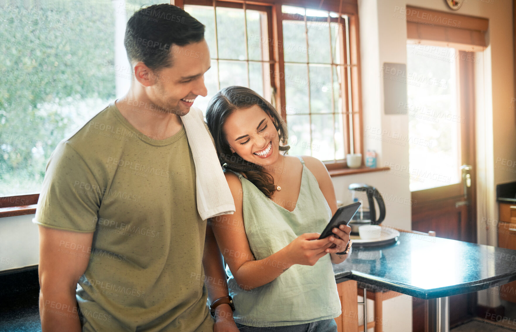 Buy stock photo Young cheerful mixed race couple using a phone together in kitchen at home. Hispanic girlfriend showing her boyfriend a funny picture on her cellphone. Man smiling at video on a phone with his wife