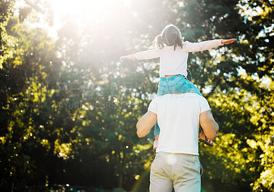 Father and playful daughter from behind having fun in sun at park with copyspace on green trees. Loving parent carrying cheerful girl on shoulders for piggyback ride with arms out pretending to fly