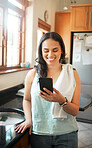 Happy mixed race woman smiling while using smart phone at home. Woman reading text message or chatting on social media while busy with chores in the kitchen 