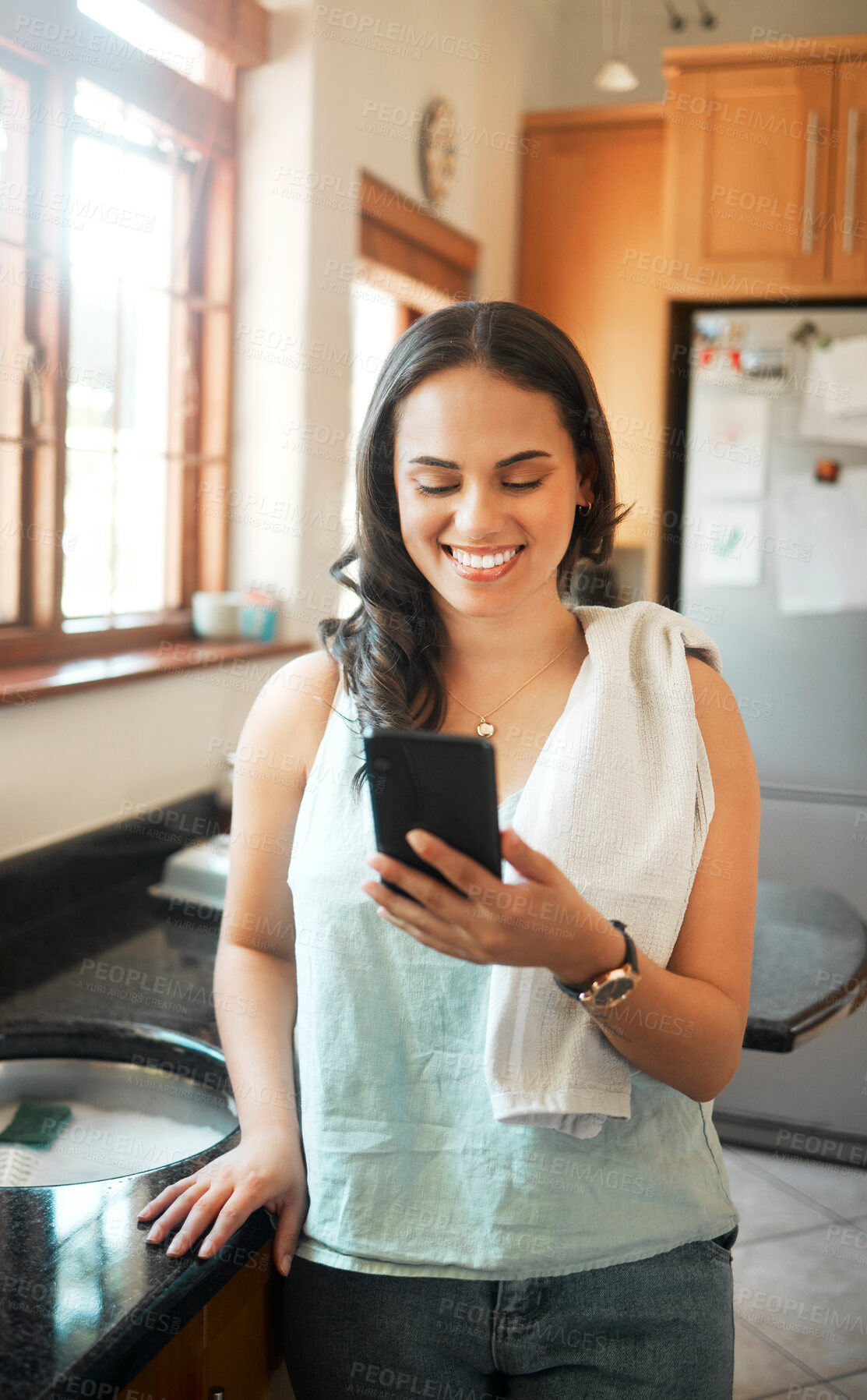 Buy stock photo Happy mixed race woman smiling while using smart phone at home. Woman reading text message or chatting on social media while busy with chores in the kitchen