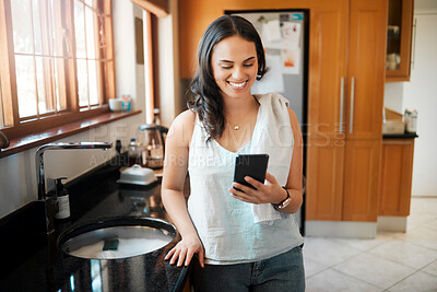Buy stock photo Happy mixed race woman smiling while using smart phone at home. Woman reading text message or chatting on social media while busy with chores in the kitchen. Young woman texting while spring cleaning