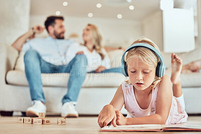Buy stock photo Little caucasian girl reading a story book while wearing headphones listening to music with her parents in the background. Child reading a fairytale and listening to music with her mom and dad at home
