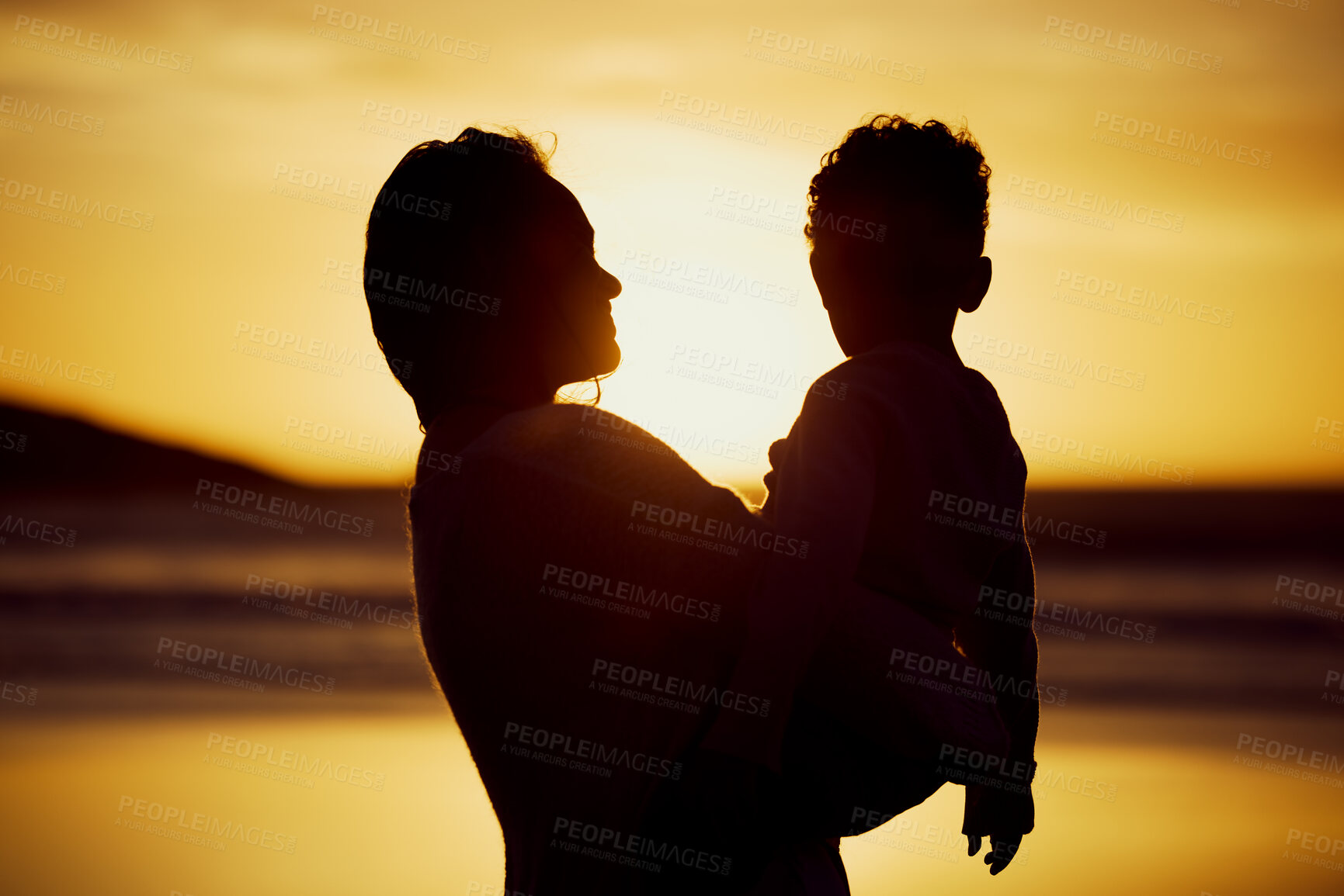 Buy stock photo Silhouette shot of a mother holding her son on the beach at sunset. Woman and kid spending time together at the beach against golden sky. Mom and son sharing a beautiful bond 