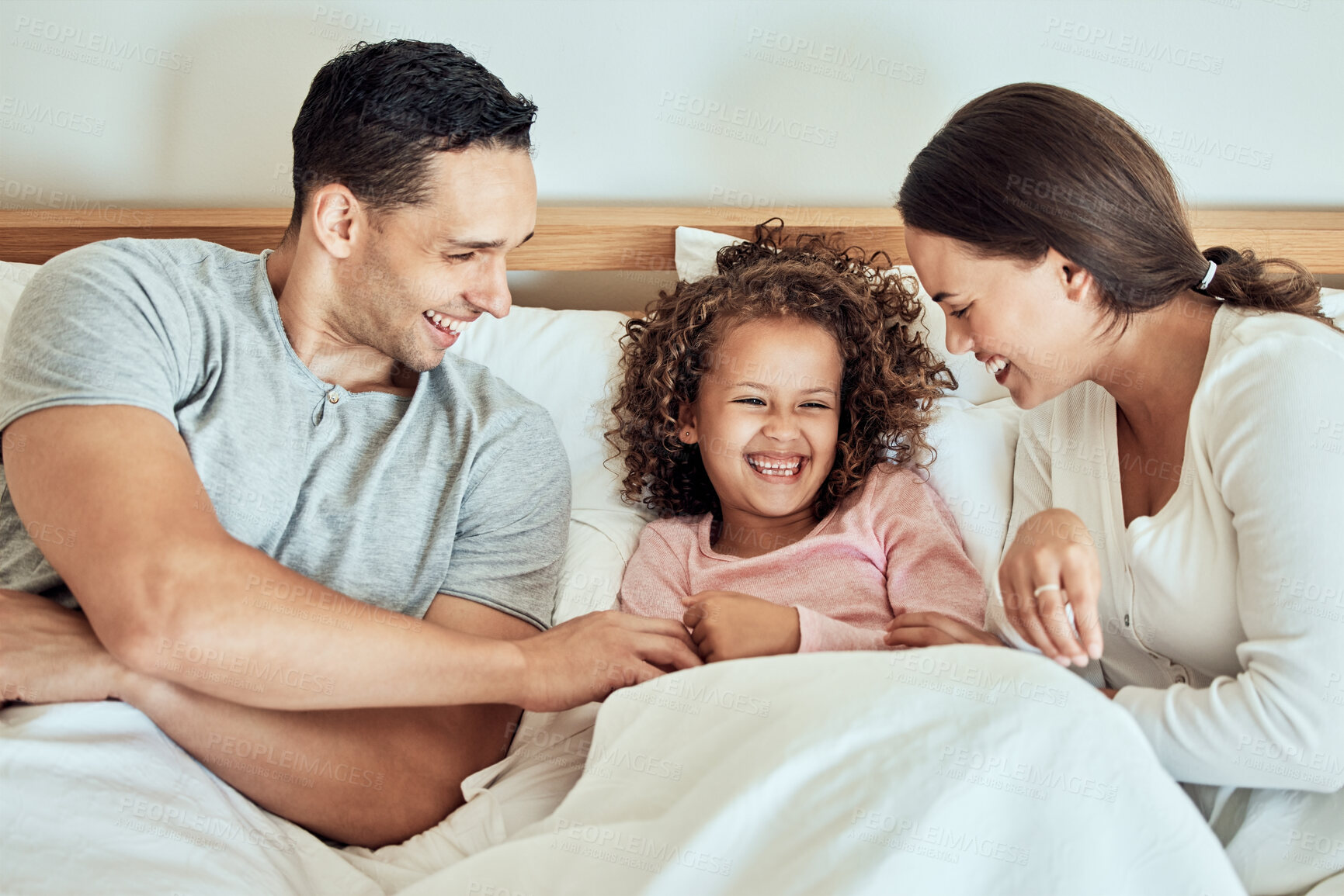 Buy stock photo Playful parents tickling their little daughter while lying in bed together. Cheerful couple bonding with their adorable little girl at home in the morning. Cute little girl laughing while playing with mom and dad. Waking up happy