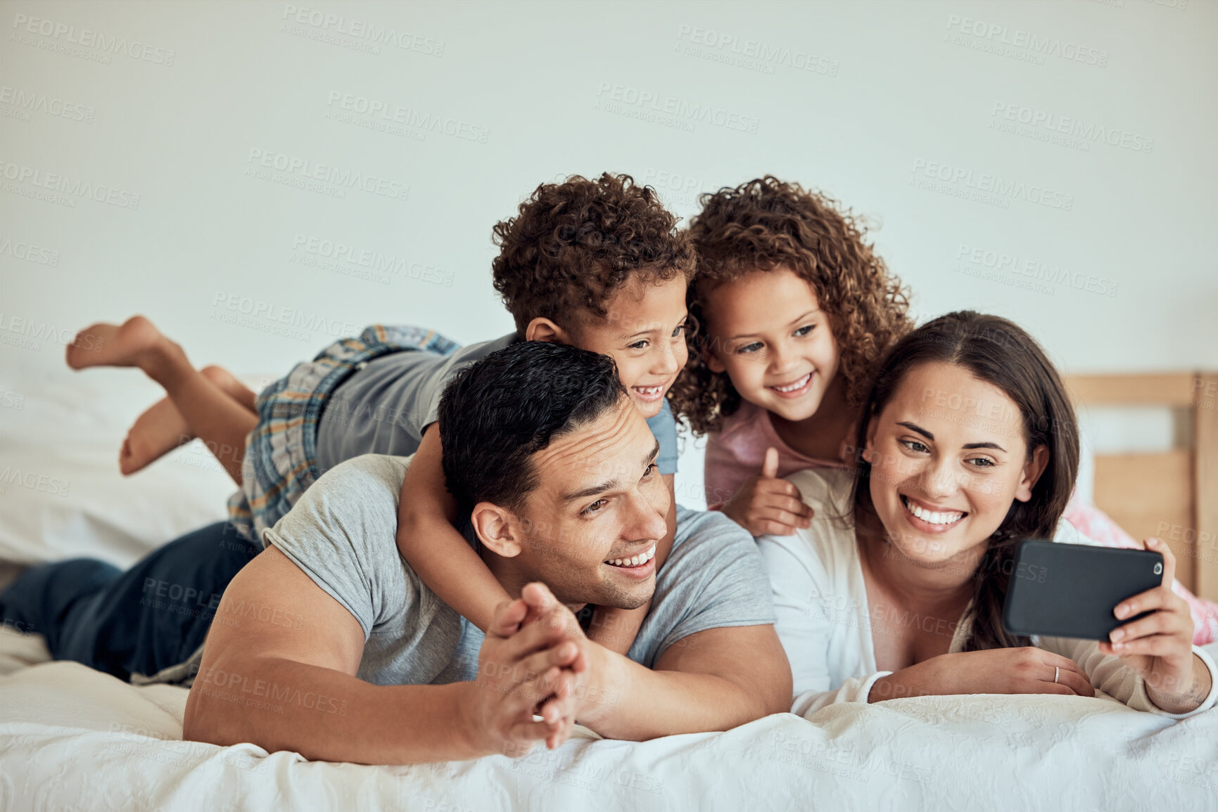 Buy stock photo Happy family with little kids taking selfies on a cellphone while lying on bed at home. Smiling couple bonding with son and daughter on a morning at home. Adorable boy and girl with mother and father