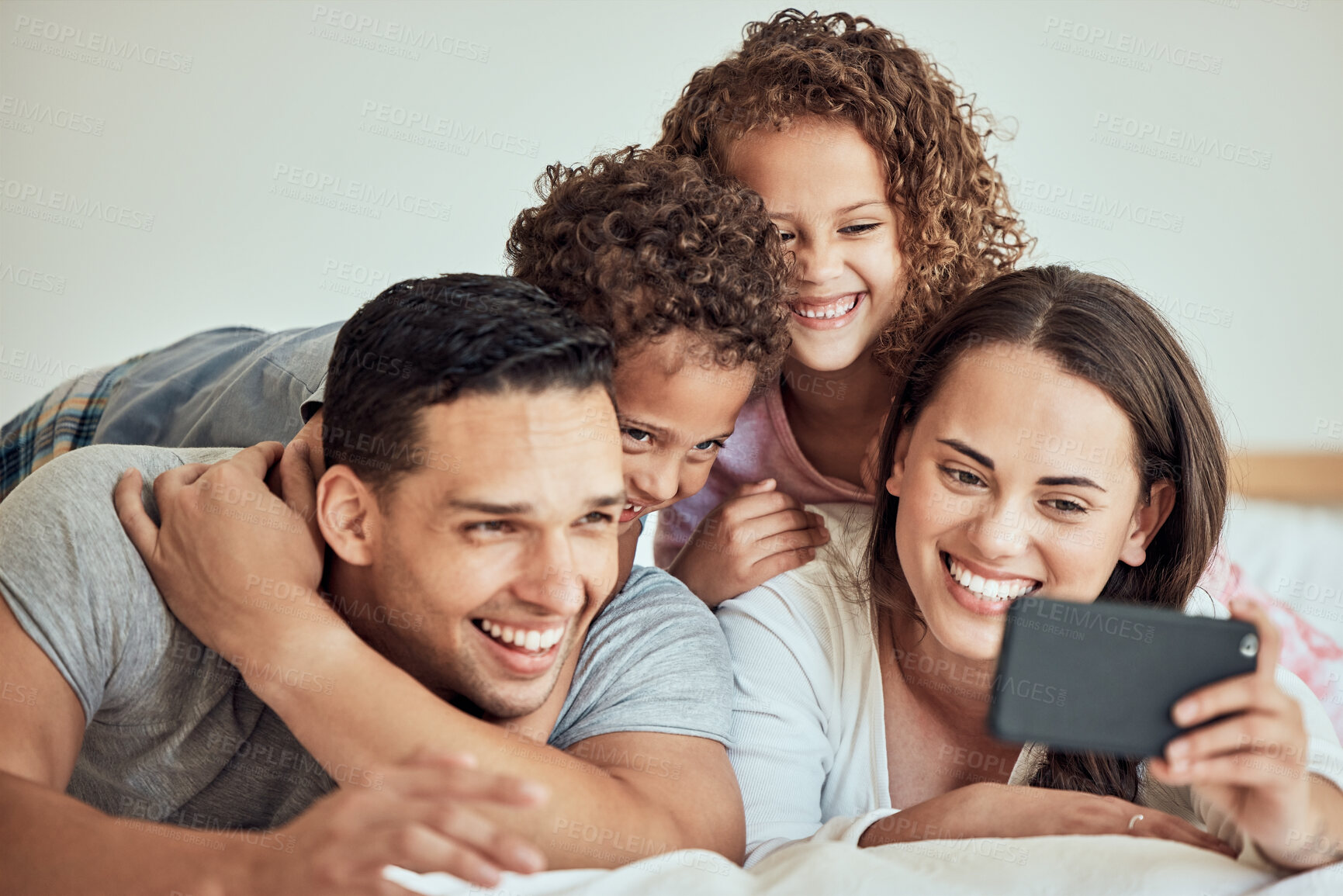 Buy stock photo Young happy mixed race family smiling for a selfie lying on a bed together at home. Hispanic woman smiling taking a photo with her children and husband with her cellphone relaxing in a bedroom