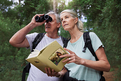 Senior caucasian couple using binoculars and a book while doing bird watching in a forest during a hike. Mature husband and wife exploring in a forest