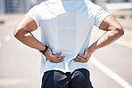 Rear view of a male athlete standing with his hands on his back suffering from sports injure during his run on a urban city road. Young sportsman experiencing back pain