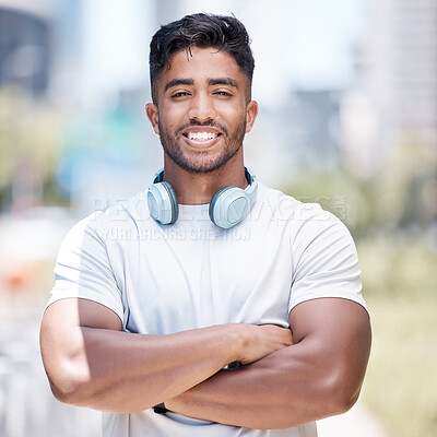 Fitness man wearing headphones around his neck while looking at the camera and smiling. Cheerful male runner standing with his arms crossed while out in the city for a workout