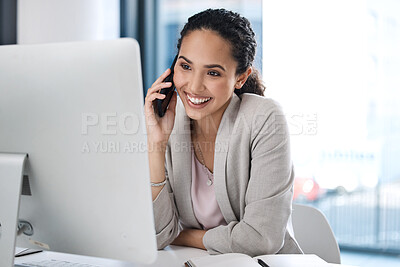 Young happy mixed race businesswoman on a call using her phone and working on a desktop computer in an office at work. One hispanic businesswoman smiling while sitting at a desk and talking on her cellphone at work
