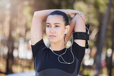 Portrait of a fit young hispanic woman wearing earphones and looking away while stretching her arms behind her head before a run. Female athlete warming up and getting ready for a cardio workout at park