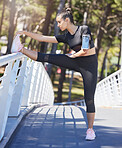 Fit young mixed race sportswoman stretching her legs on a bridge in park on a sunny day. Female athlete wearing phone armband while doing warmup exercise in nature