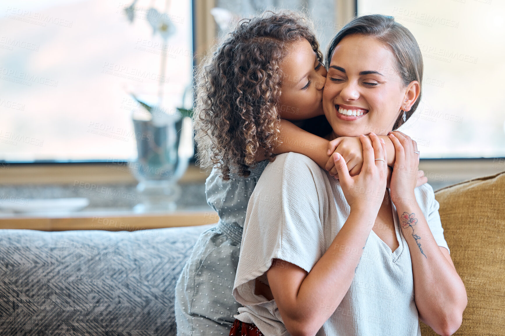 Buy stock photo Little daughter embracing her mother from behind and gently kissing her on the cheek showing love and affection while sitting on the couch at home. Sweet moment between mother and child on mothers day