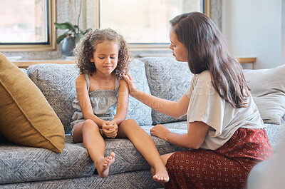 Sad little girl sitting on couch while mother tries to talk to her. Loving caring mother trying to communicate with upset daughter. Young hispanic mother asking little girl whats wrong while trying to comfort her and show support