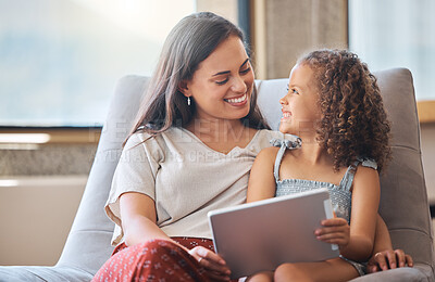 Happy little girl and loving mother looking at each other while sitting together on couch and using digital tablet to watch a movie or do a video call with family. Parent sitting with child while enjoying some online entertainment