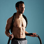 Handsome young hispanic man training with battle ropes in studio isolated against a blue background. Mixed race shirtless male athlete exercising or working out to increase his strength and fitness