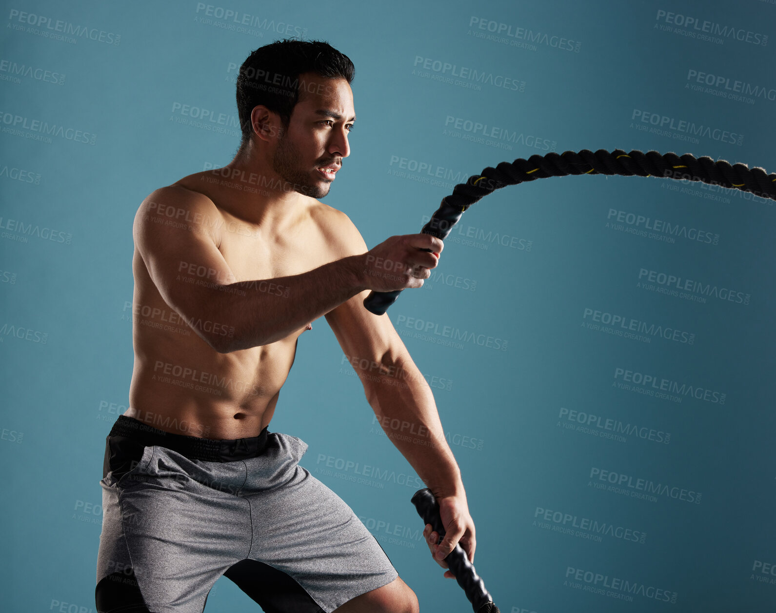 Buy stock photo Handsome young hispanic man training with battle ropes in studio isolated against a blue background. Mixed race shirtless male athlete exercising or working out to increase his strength and fitness