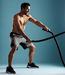 Handsome young hispanic man training with battle ropes in studio isolated against a blue background. Mixed race shirtless male athlete exercising or working out to increase his strength and fitness