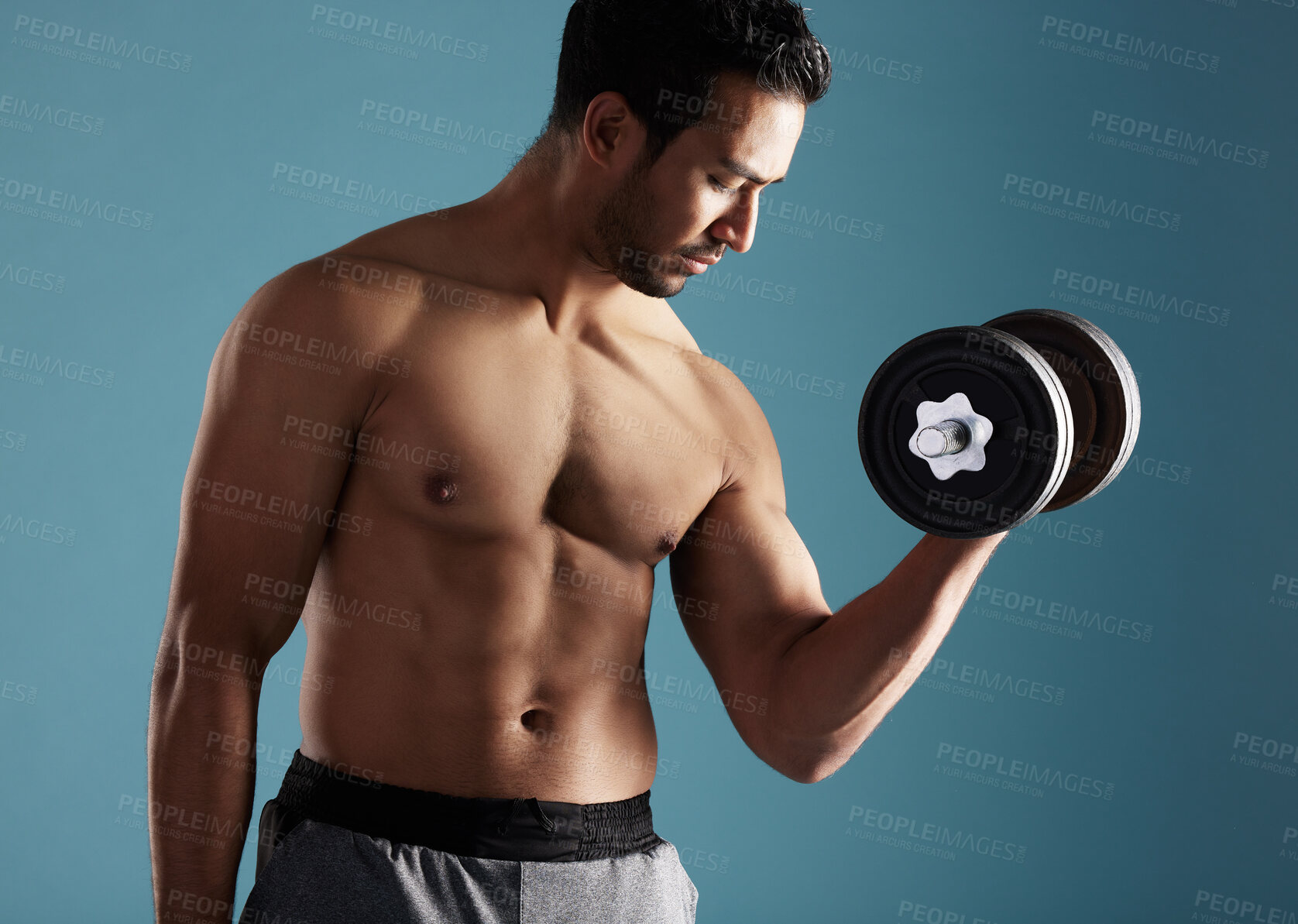 Buy stock photo Handsome young hispanic man training with dumbbells in studio isolated against a blue background. Mixed race shirtless male athlete exercising or working out to increase his strength and fitness