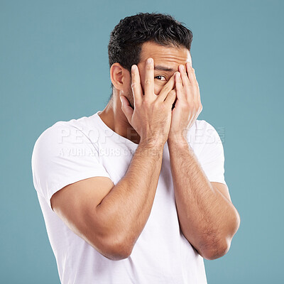 Buy stock photo Handsome young mixed race man peaking through his fingers and covering his face with his hands while standing in studio isolated against a blue background. Hispanic male looking shy and hiding away
