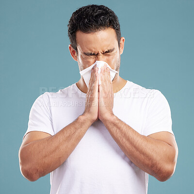 Handsome young mixed race man blowing his nose while standing in studio isolated against a blue background. Hispanic male suffering from cold, flu, sinus, hayfever or corona and using a facial tissue