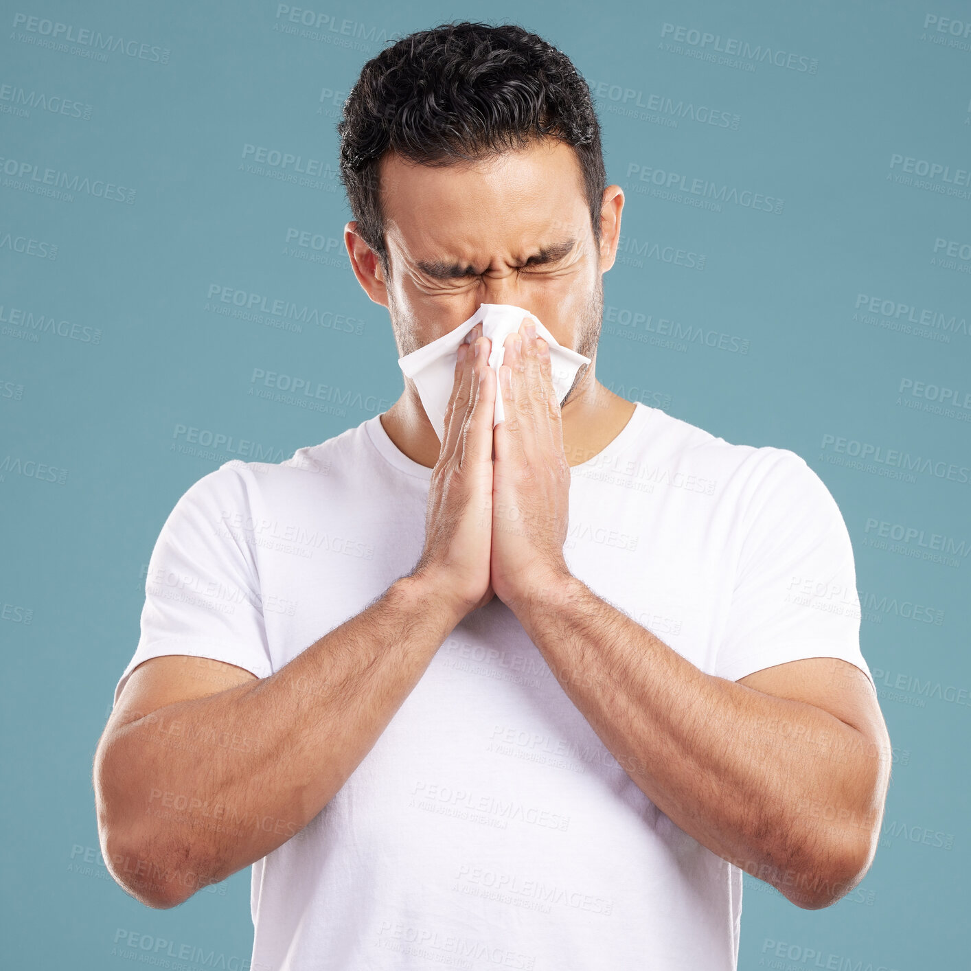 Buy stock photo Handsome young mixed race man blowing his nose while standing in studio isolated against a blue background. Hispanic male suffering from cold, flu, sinus, hayfever or corona and using a facial tissue