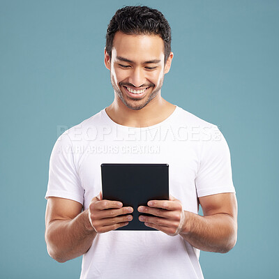 Buy stock photo Handsome young mixed race man using his tablet while standing in studio isolated against a blue background. Hispanic male sending an online message, using the internet or browsing social media
