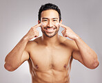 Handsome young mixed race man shirtless in studio isolated against a grey background. Hispanic male applying moisturizer lotion to his face. Looking confident and happy with his daily skincare regime