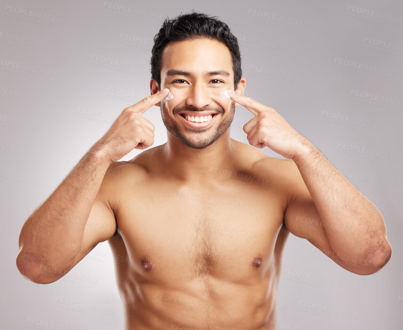 Buy stock photo Handsome young mixed race man shirtless in studio isolated against a grey background. Hispanic male applying moisturizer lotion to his face. Looking confident and happy with his daily skincare regime