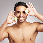 Handsome young mixed race man posing with cucumber slices isolated in studio against a grey background. His skincare regime keeps him fresh. Eating healthy vitamins and nutrients for firm skin