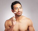 Handsome young mixed race man posing shirtless in studio isolated against a grey background. Hispanic male using a dermal face roller to massage and relax his skin. All part of his skincare regime