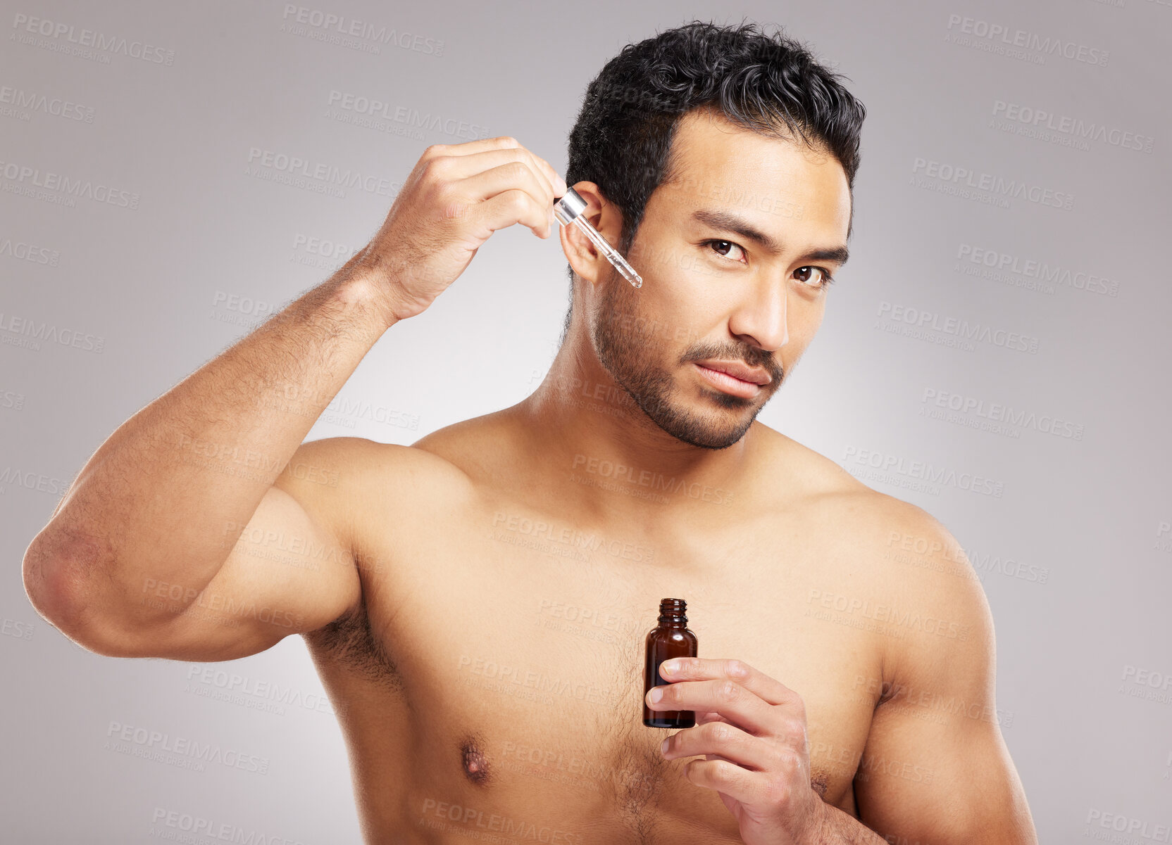 Buy stock photo Handsome young mixed race man posing shirtless in studio isolated against a grey background. Hispanic male applying antiaging serum to his face using a dropper. All part of his skincare regime