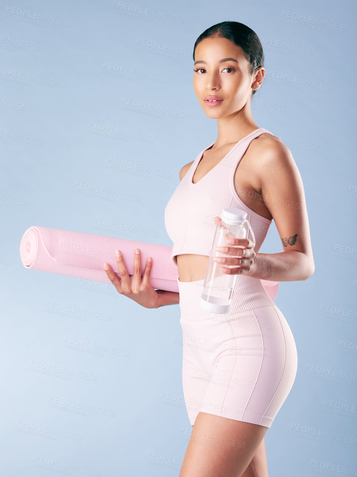 Buy stock photo Mixed race fitness woman standing with her yoga mat and water bottle in studio against a blue background. Beautiful young hispanic female athlete exercising or working out. Health and fitness