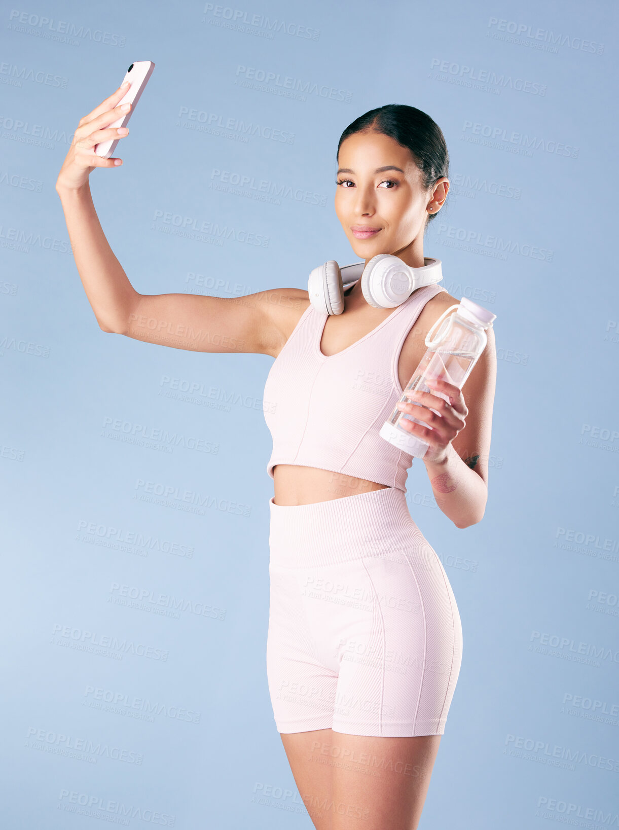 Buy stock photo Mixed race fitness woman posing with her phone in studio against a blue background. Young hispanic female athlete taking selfie pictures with her smartphone to track her personal fitness growth