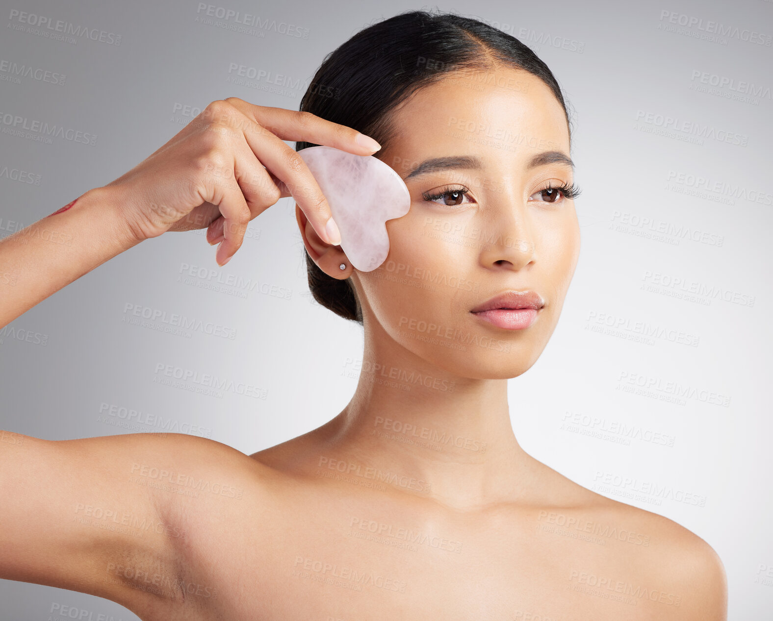 Buy stock photo Studio closeup of a beautiful mixed race woman using a rose quartz gua sha to reduce wrinkles and pufiness. Young hispanic woman using anti ageing tool against grey copyspace background