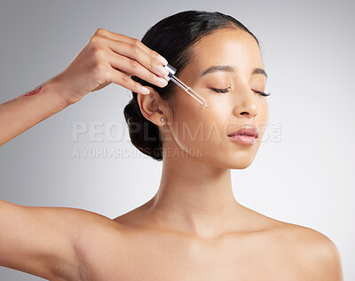 A beautiful mixed race woman applying a soothing face serum to her radiant smooth face while posing against a grey background. Hispanic woman with flawless skin using essential oil