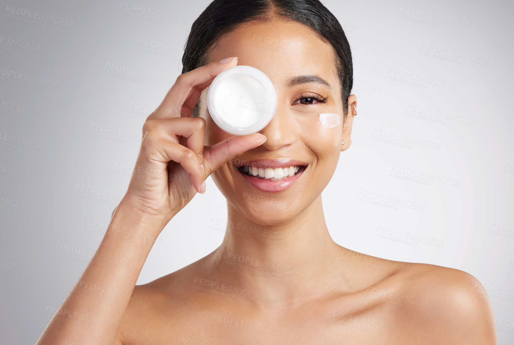 Buy stock photo Studio Portrait of a beautiful smiling mixed race woman applying cream to her face. Hispanic model with glowing skin holding moisturiser against a grey copyspace background