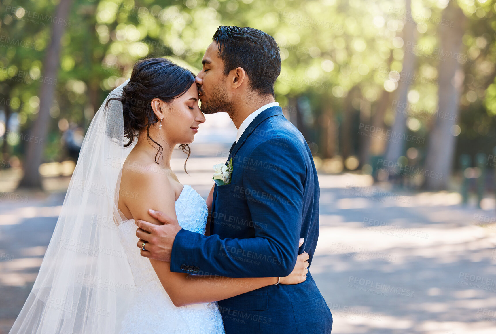 Buy stock photo Loving husband kissing his beautiful bride on the forehead. Happy in love newlywed couple standing together sharing romantic moment outdoors