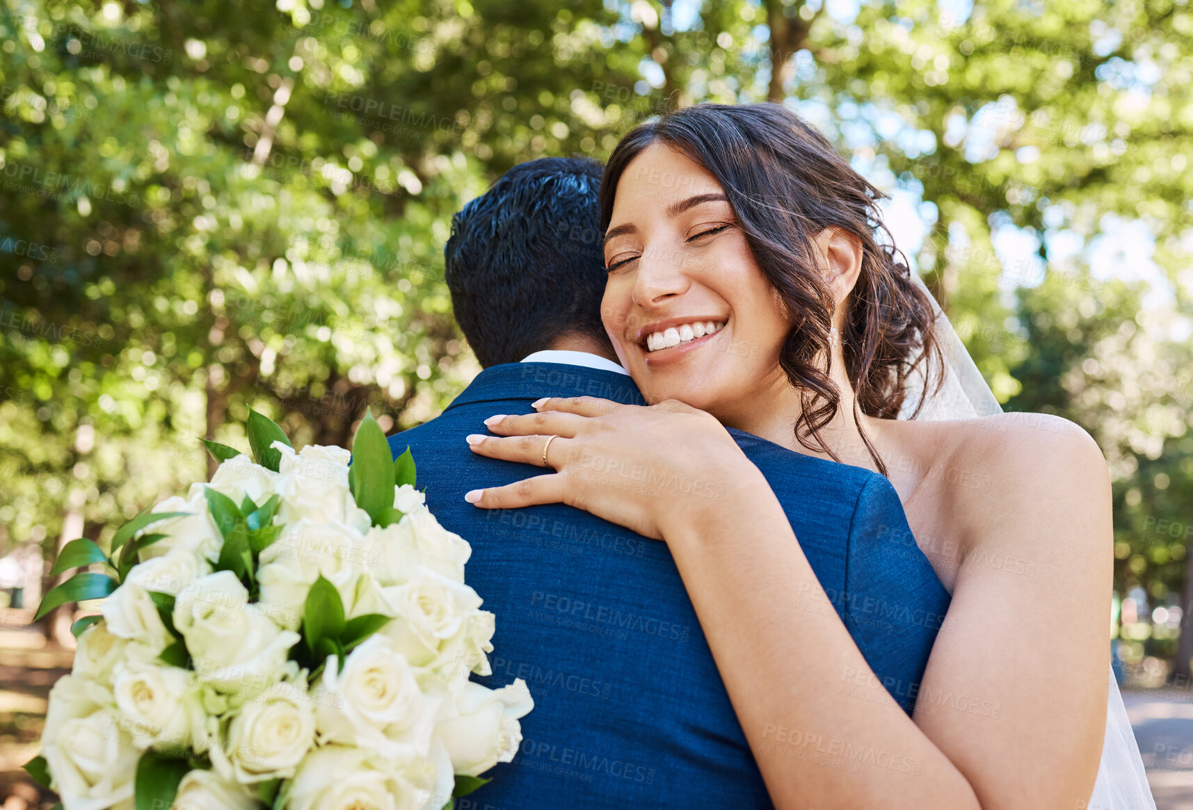 Buy stock photo Beautiful joyful bride holding bouquet and embracing her husband. Happy newlywed couple hugging and looking happy on their wedding day against nature background