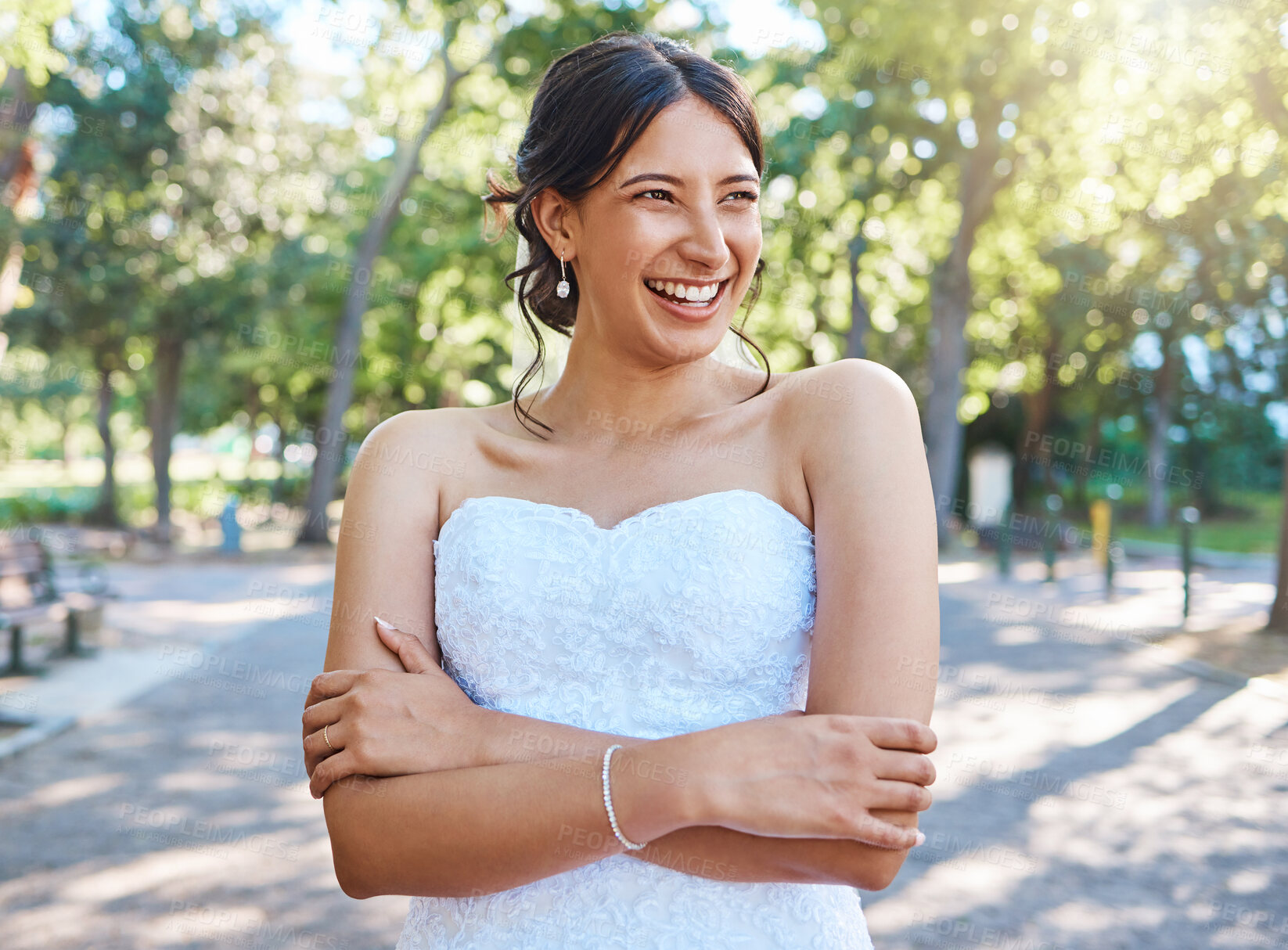 Buy stock photo Beautiful mixed race bride wearing a white strapless dress while standing with her arms crossed in nature on a sunny day. Carefree bride smiling and dreaming about her her future as a wife
