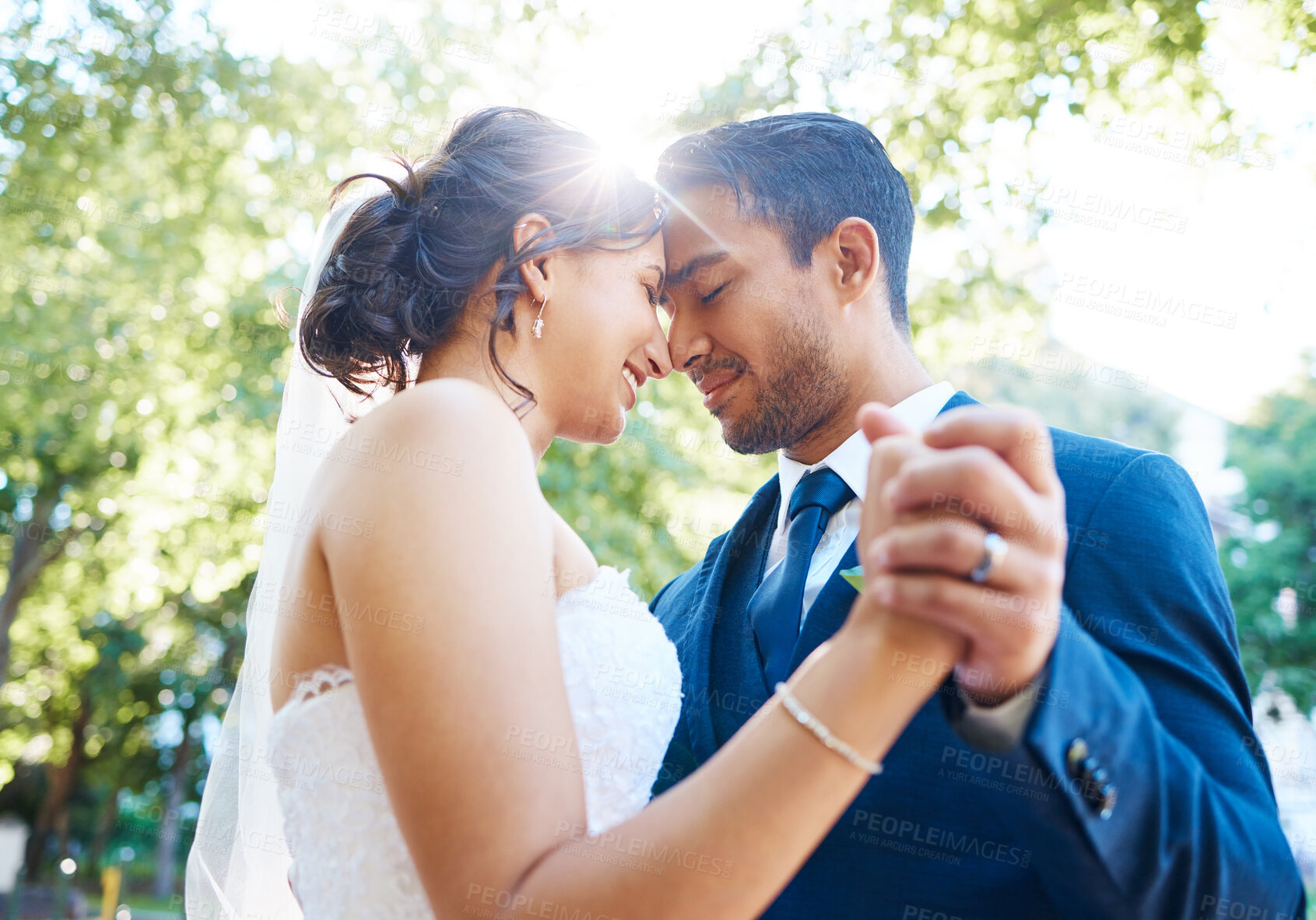 Buy stock photo Bride and groom holding hands and touching foreheads while dancing outside. Mixed race newlyweds enjoying romantic moments on their wedding day. Happy young romantic couple sharing their first wedding dance