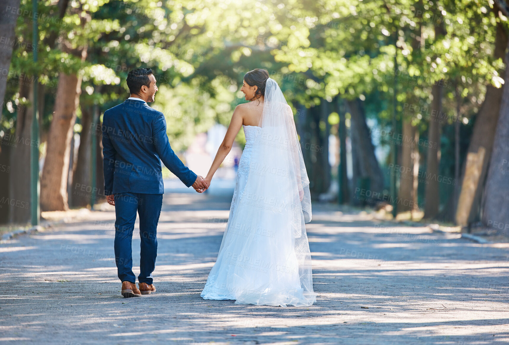 Buy stock photo Rear view shot of newlywed couple holding hands and walking through park. Bride and groom walking away after their wedding reception. Living happily ever after