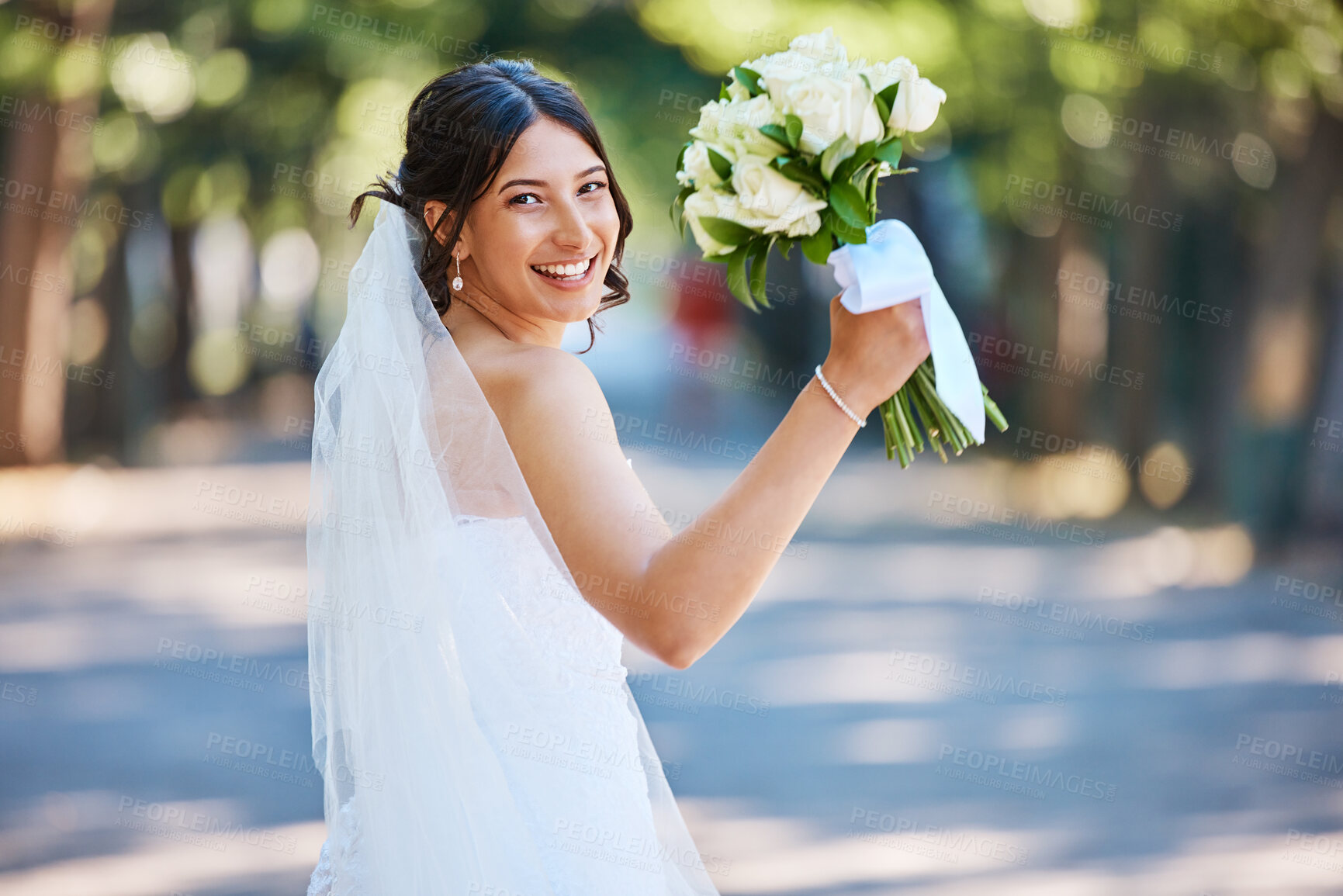 Buy stock photo Portrait of a beautiful mixed race bride looking back over her shoulder and holding up her bouquet. Stunning bride walking in a park