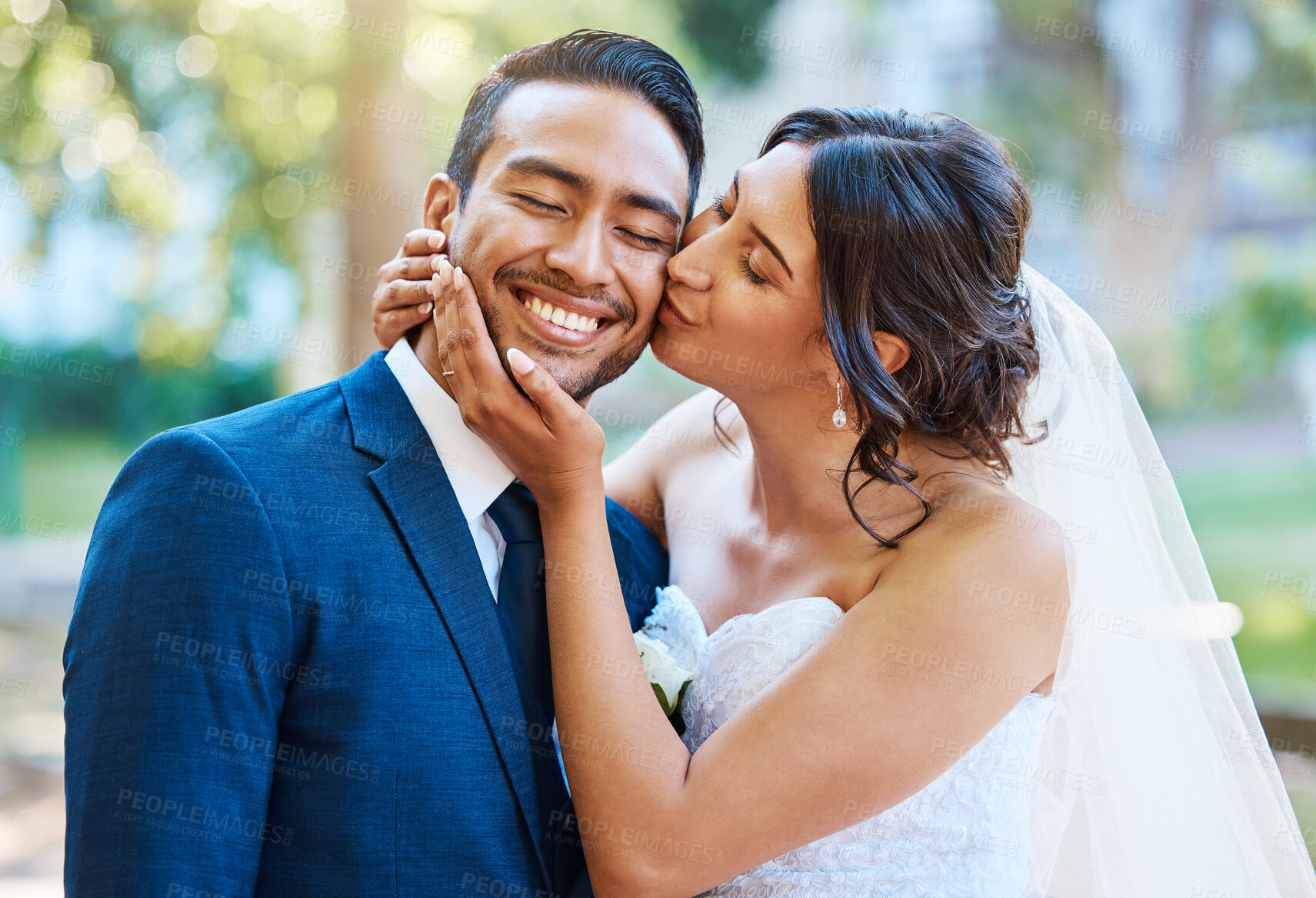 Buy stock photo Beautiful bride kissing her groom on the cheek. Happy loving newlyweds enjoying romantic moments on their wedding day in nature