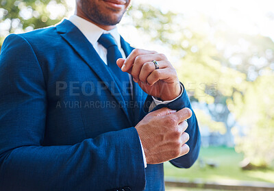 Close up of a groom wearing a blue suit with a white shirt and tie. Bridegroom adjusting his cufflinks and wearing a wedding band while standing outdoors on a sunny day