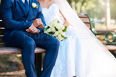 Closeup of a bride and groom sitting on a park bench together. Man wearing suit and woman wearing wedding dress and holding bouquet while sitting outdoors