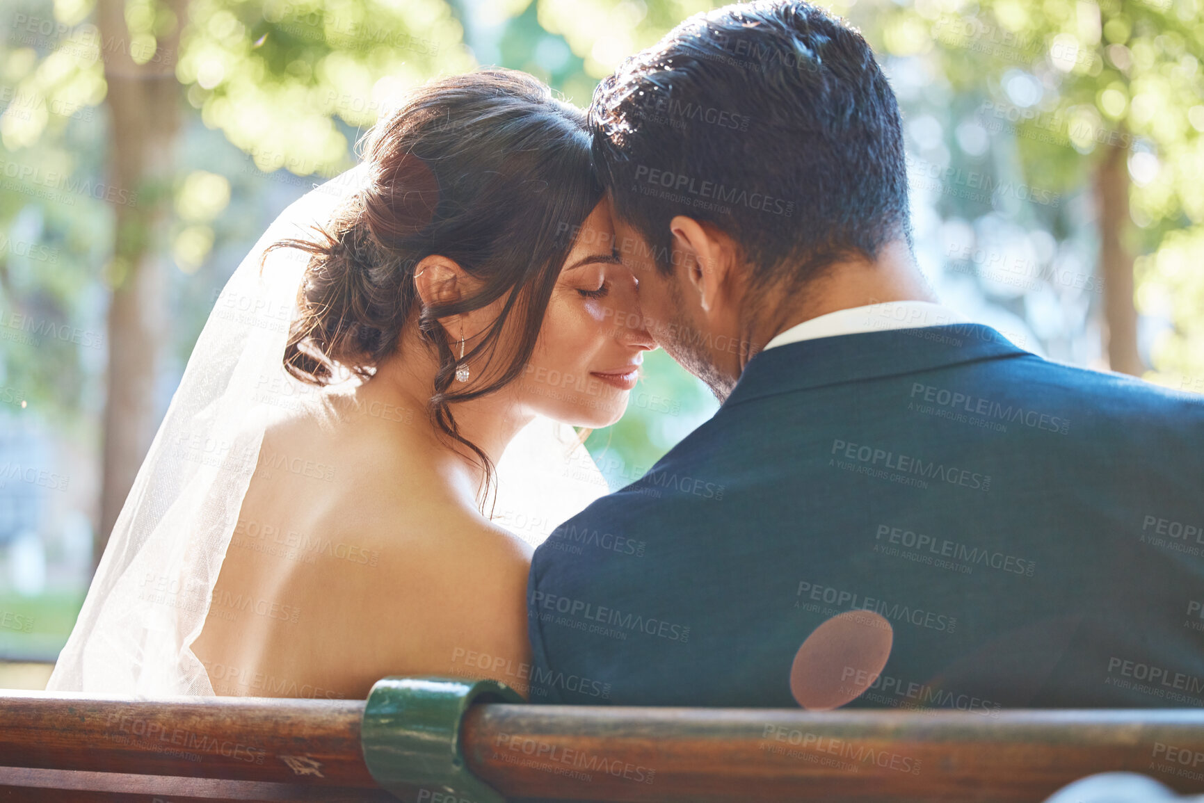 Buy stock photo Rear view of loving couple enjoying romantic moments on their wedding day. Newlywed couple touching foreheads while leaning against each other while sitting on a park bench