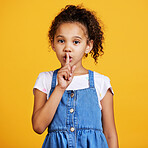 Studio portrait mixed race girl standing with her finger on her lips isolated against a yellow background. Cute hispanic child posing inside. Happy and cute kid saying quiet, whisper or keep a secret
