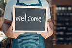 Closeup of one mixed race man holding a chalkboard with the word "welcome" written on in a cafe or store. Hands of guy holding sign to greet customers at the entrance of an open shop